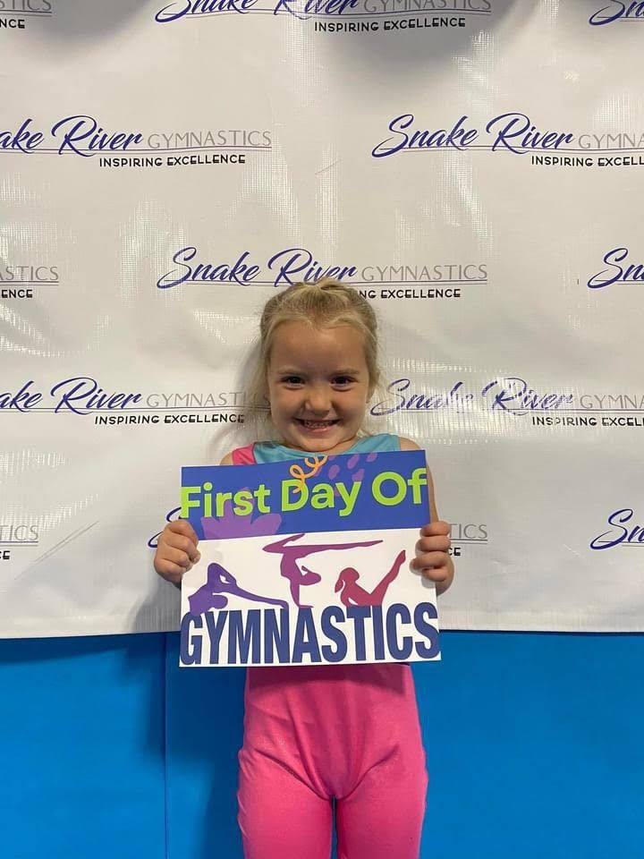 A smiling girl holding a First Day of Gymnastics sign in front of a Snake River Gymnastics backdrop.