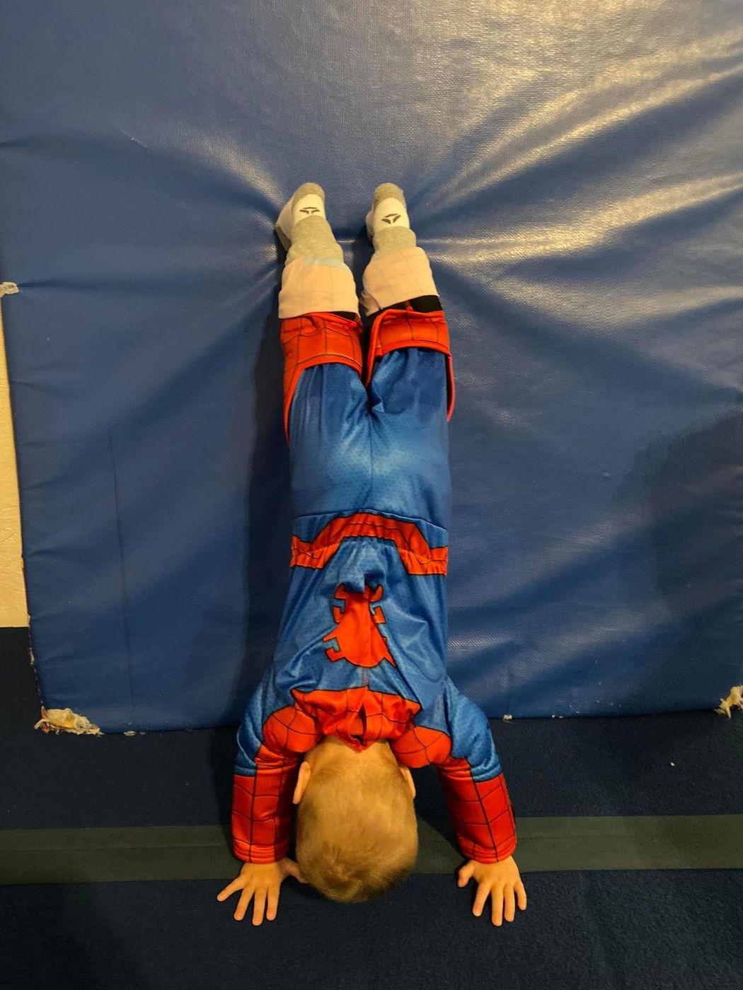 A child in a Spiderman costume doing a headstand against a blue mat.
