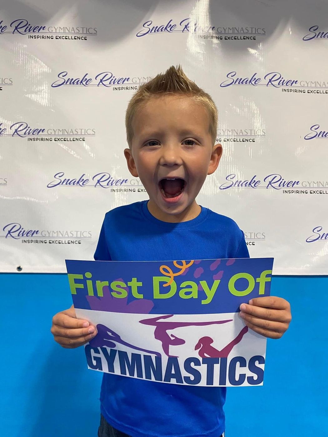 An excited boy holding a First Day of Gymnastics sign in front of a Snake River Gymnastics backdrop.