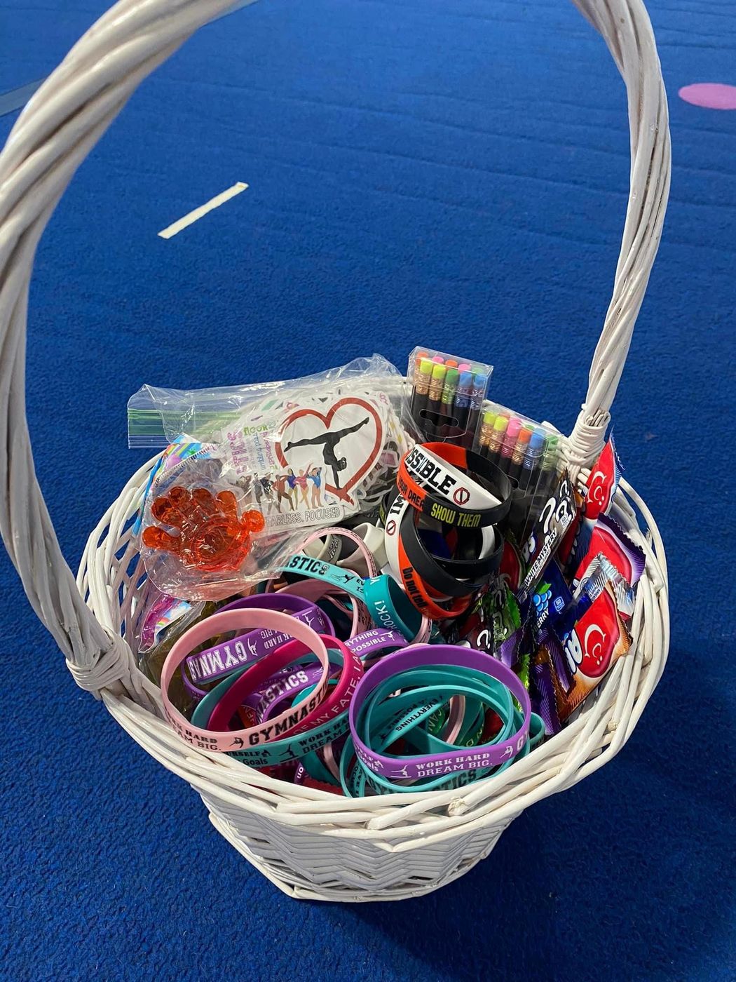 A white basket filled with gymnastics-themed bracelets, stickers and prizes on a blue mat.