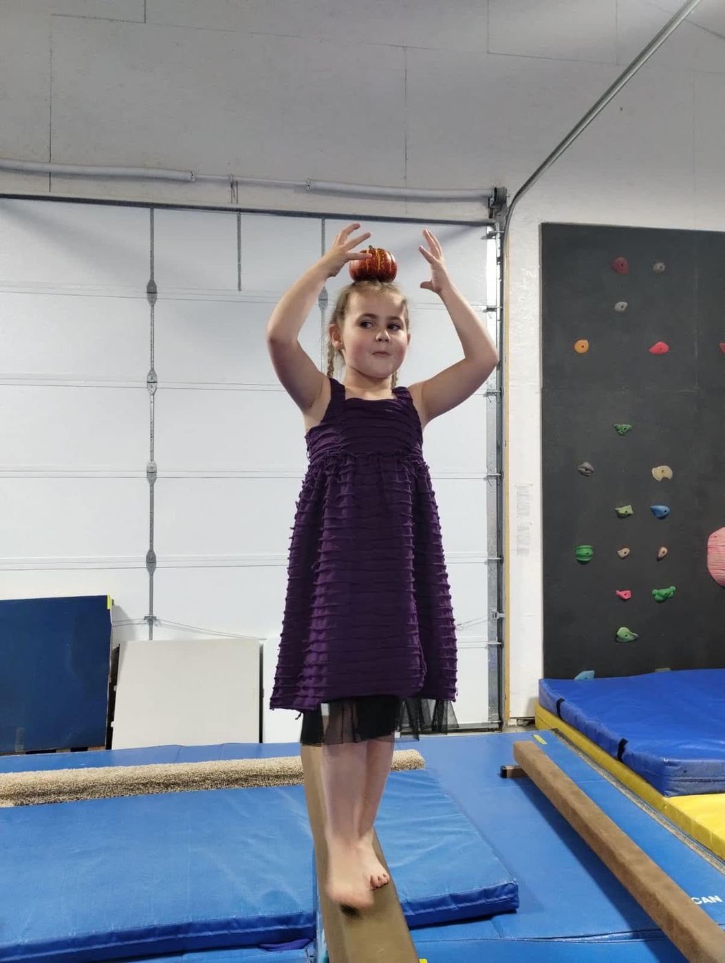 A young girl in a purple dress balancing a beanbag on her head while walking the beam.