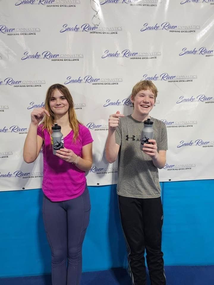 Two teenagers holding Snake River Gymnastics water bottles in front of the backdrop.