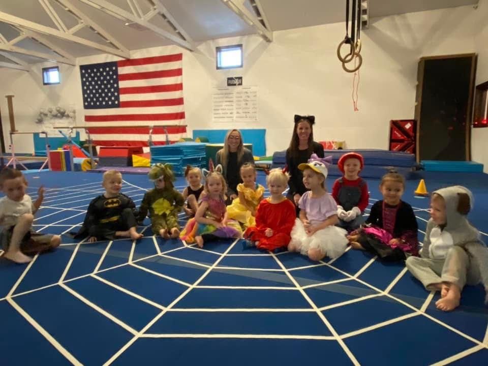 Young children in Halloween costumes seated with two coaches on a spider-web themed mat.