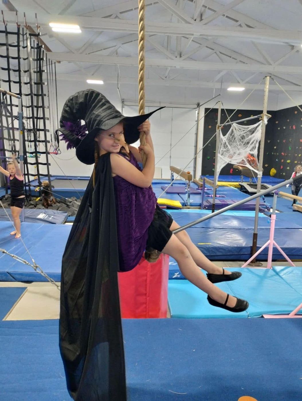 A girl in a witch costume swinging on a rope during a Halloween gym event.