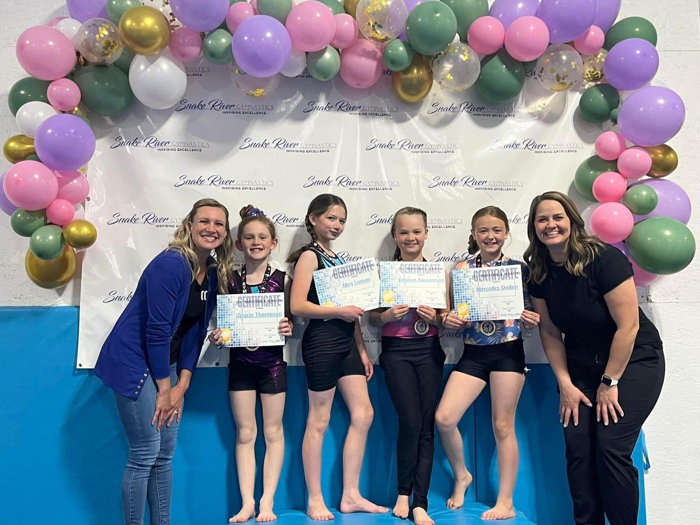 Two coaches posing with four gymnasts holding certificates under a balloon arch and Snake River Gymnastics banner.
