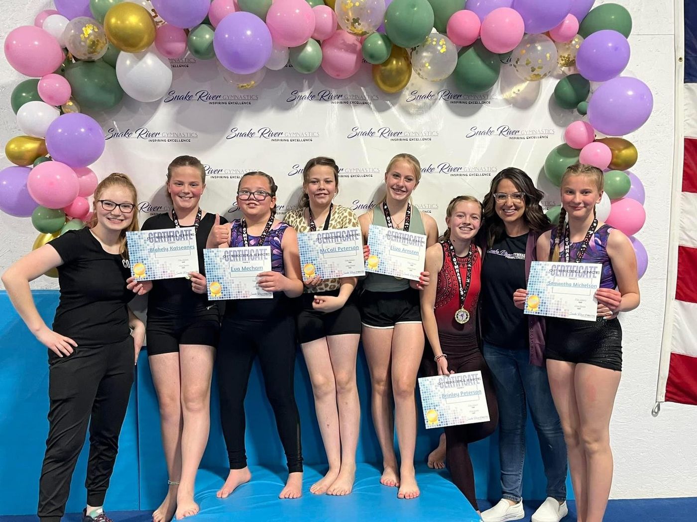 A group of teen gymnasts and coaches holding certificates and medals under a balloon arch.