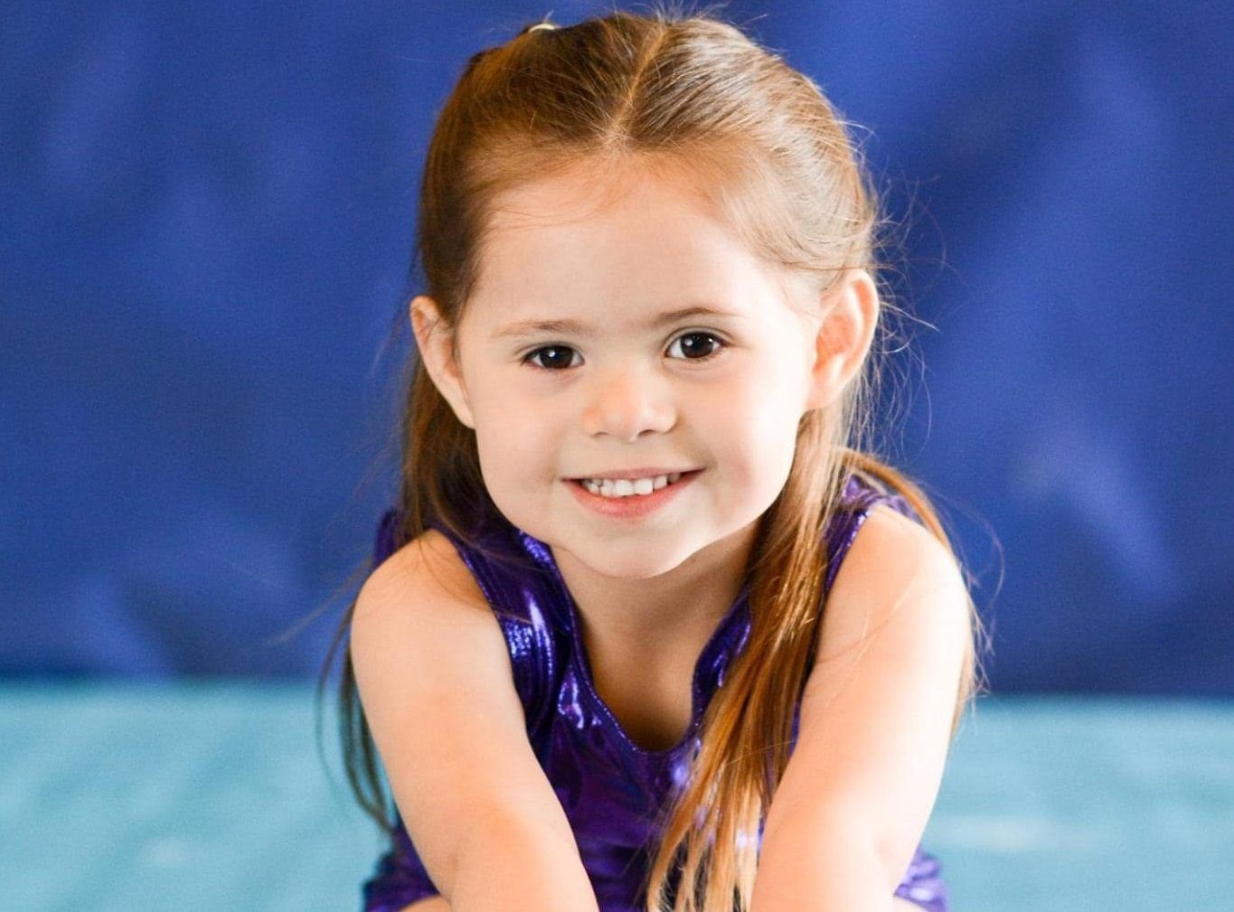 A young girl in a purple leotard smiles while leaning forward on a blue gymnastics mat.