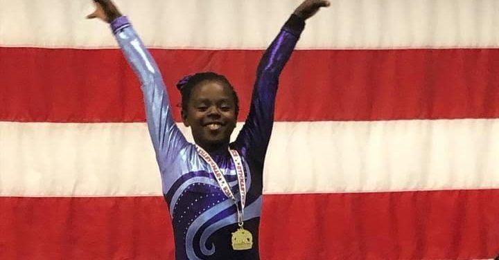 A young gymnast in a sparkly purple leotard celebrates on a podium with a gold medal in front of an American flag.