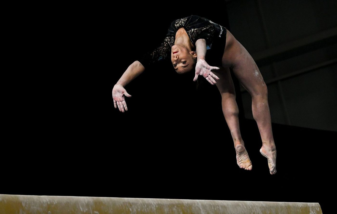 A gymnast performs an aerial flip on the balance beam against a dark background.