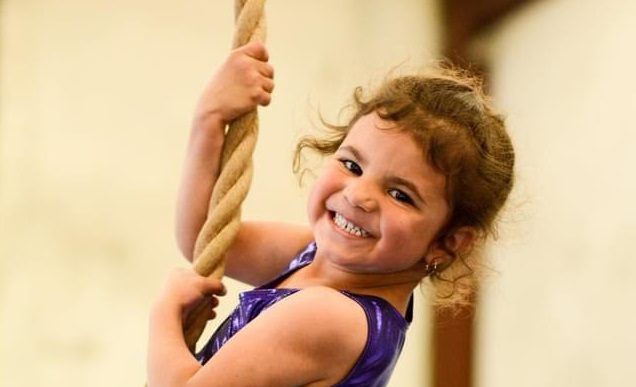A smiling young girl in a purple leotard climbs a rope in the gym.