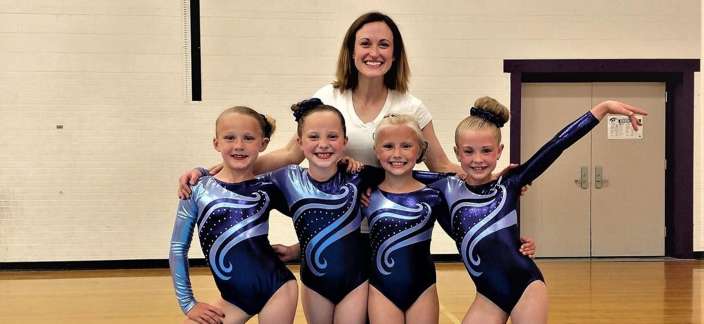 A coach in a white shirt poses with four young gymnasts in matching blue sparkly leotards in the gym.