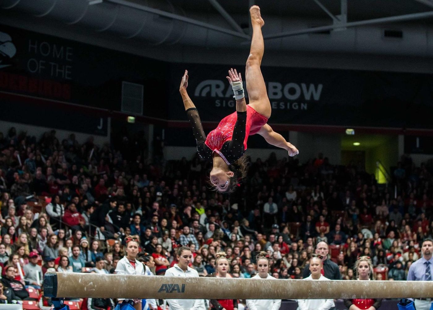 A collegiate gymnast performing a mid-air flip on the balance beam in front of a large crowd.