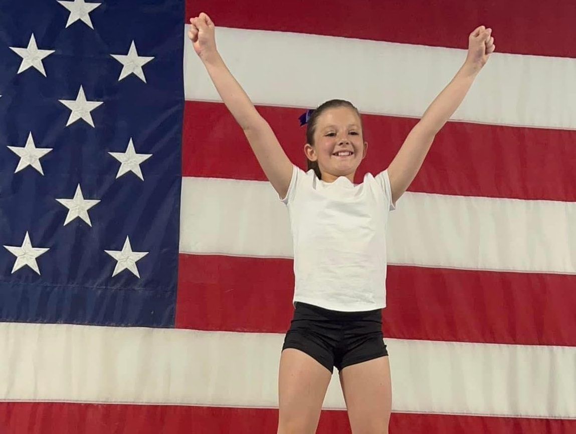 A young gymnast in a white shirt and black shorts raises her arms in a finishing pose in front of an American flag.