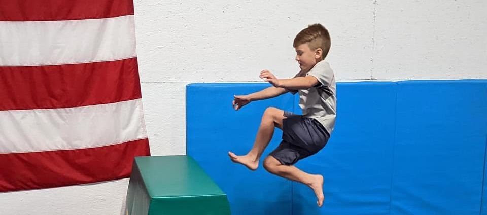 A young boy leaps mid-air toward a blue gym mat beside an American flag.
