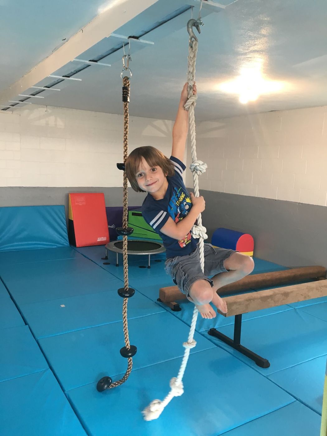 A young boy hanging from a knotted climbing rope in the parkour training room.