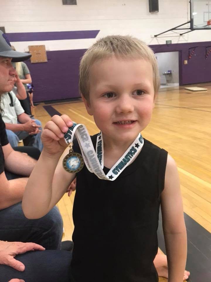 A young boy proudly holding up a gymnastics medal in a gymnasium.
