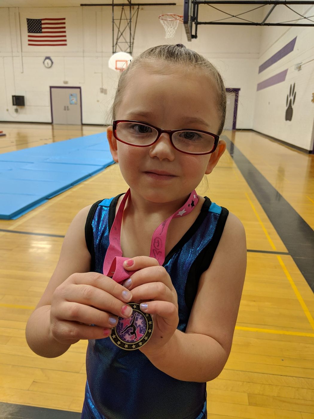 A young girl with red glasses holding up a gymnastics medal in a gymnasium.