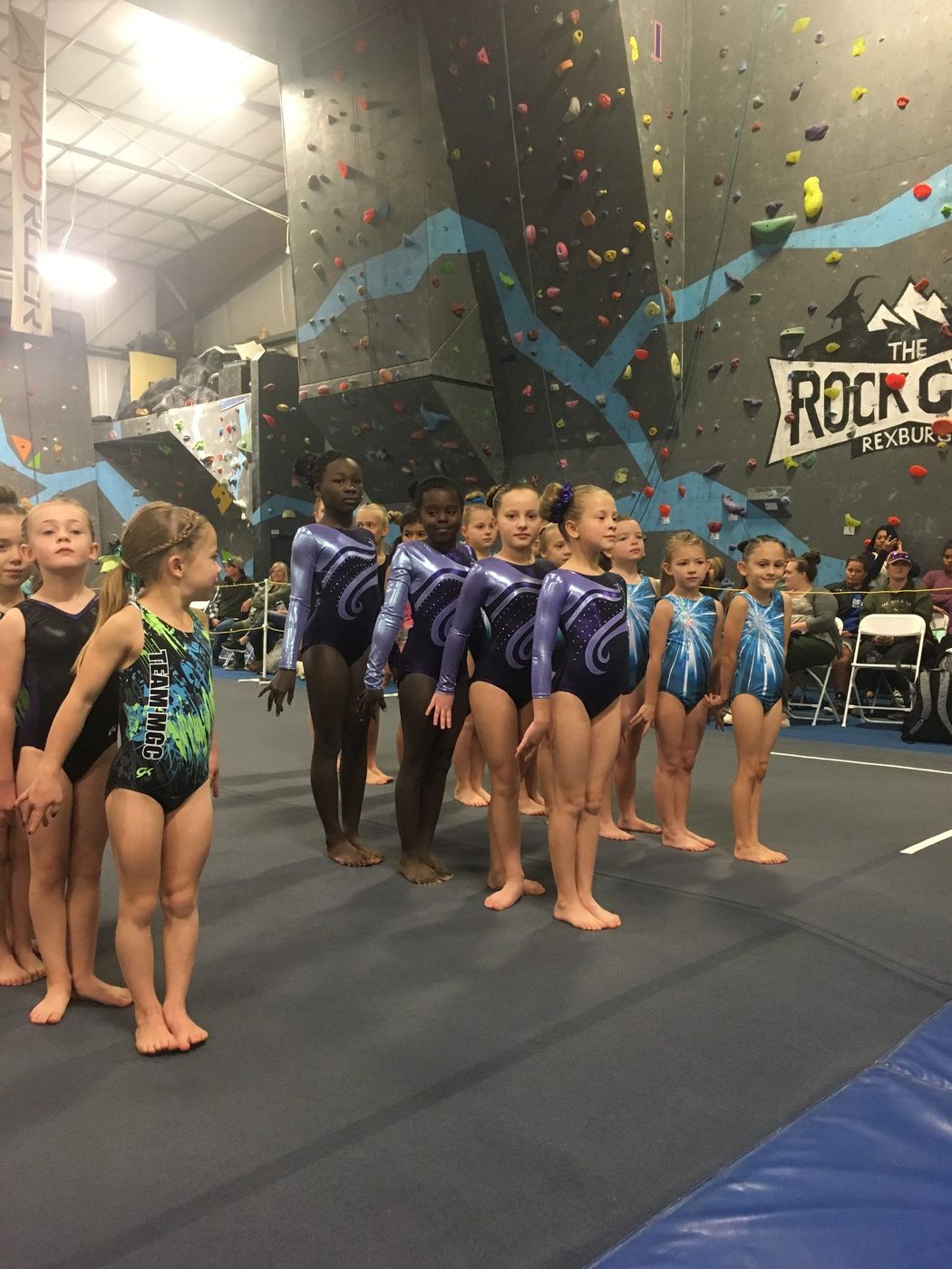A line of young gymnasts in leotards standing at attention at a meet in front of a climbing wall.