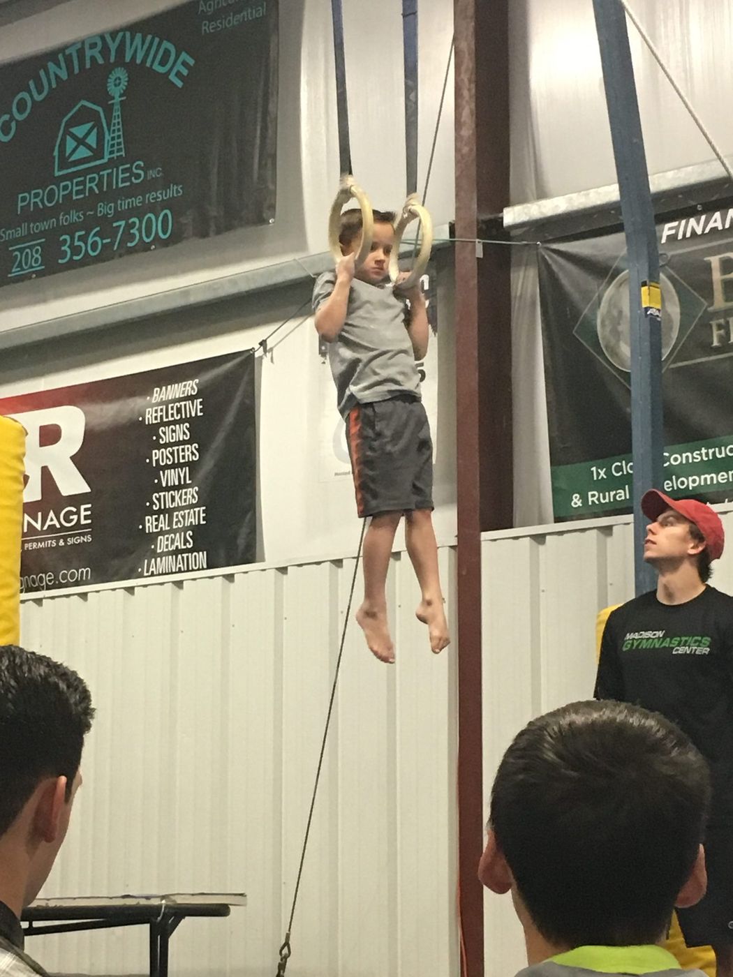 A boy hanging from the gymnastic rings at a competition while a coach watches.