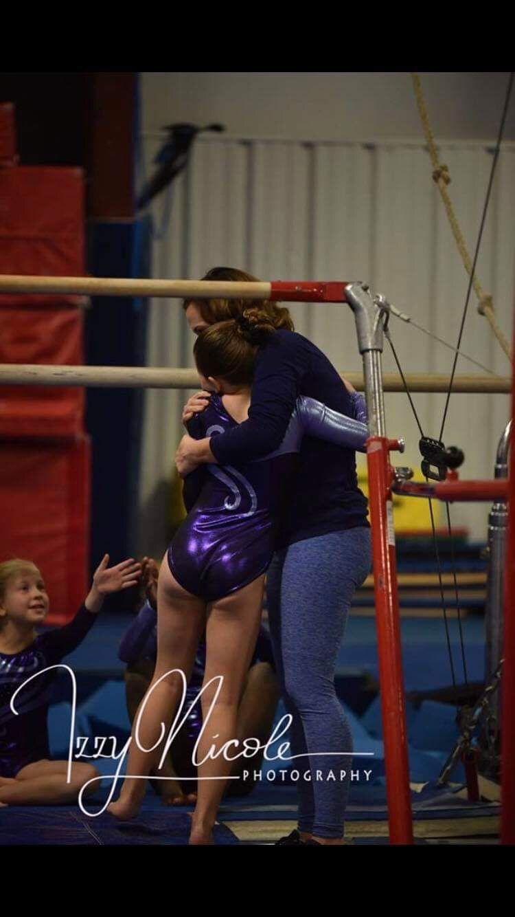 A coach hugging a young gymnast in a purple leotard beside the uneven bars.