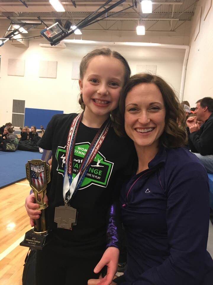 A young gymnast holding a 2nd place trophy poses with her smiling coach at a meet.