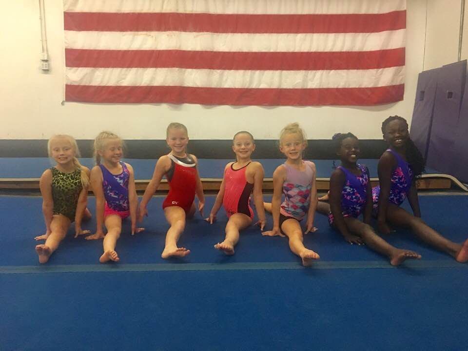 Seven young gymnasts in leotards sit in a row on a blue mat in front of an American flag.