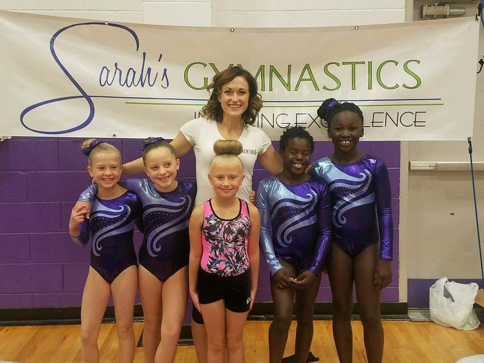 A coach poses with five young gymnasts in matching purple leotards under a Sarah's Gymnastics banner.