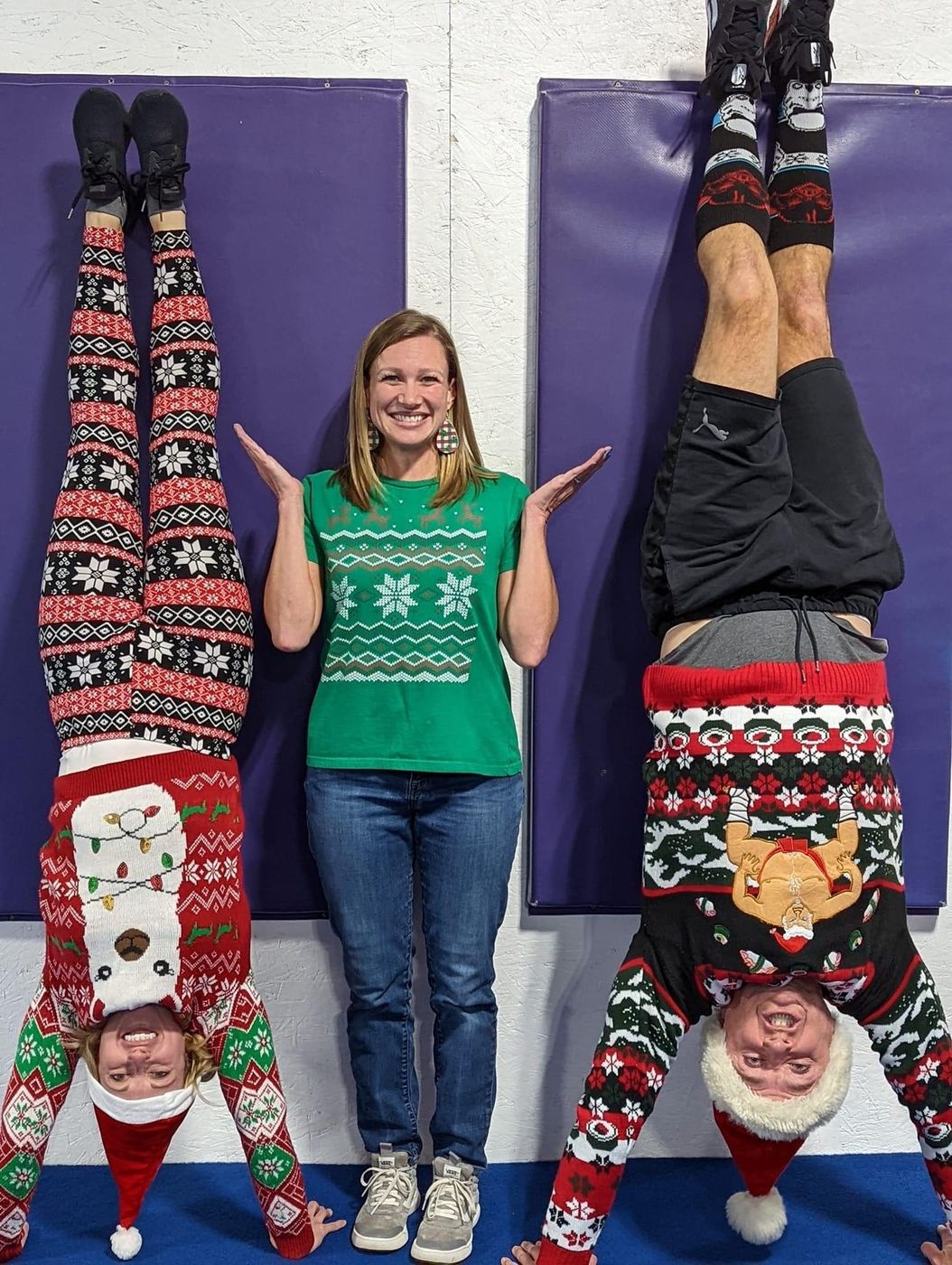 A coach in a green sweater poses between two gymnasts doing handstands in festive holiday sweaters against a purple wall.