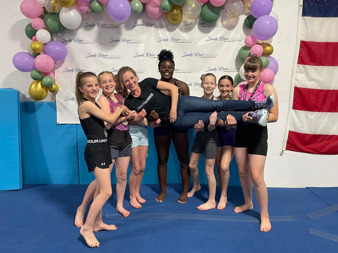 A team of young gymnasts lift one girl horizontally while posing in front of a balloon-decorated Snake River backdrop.