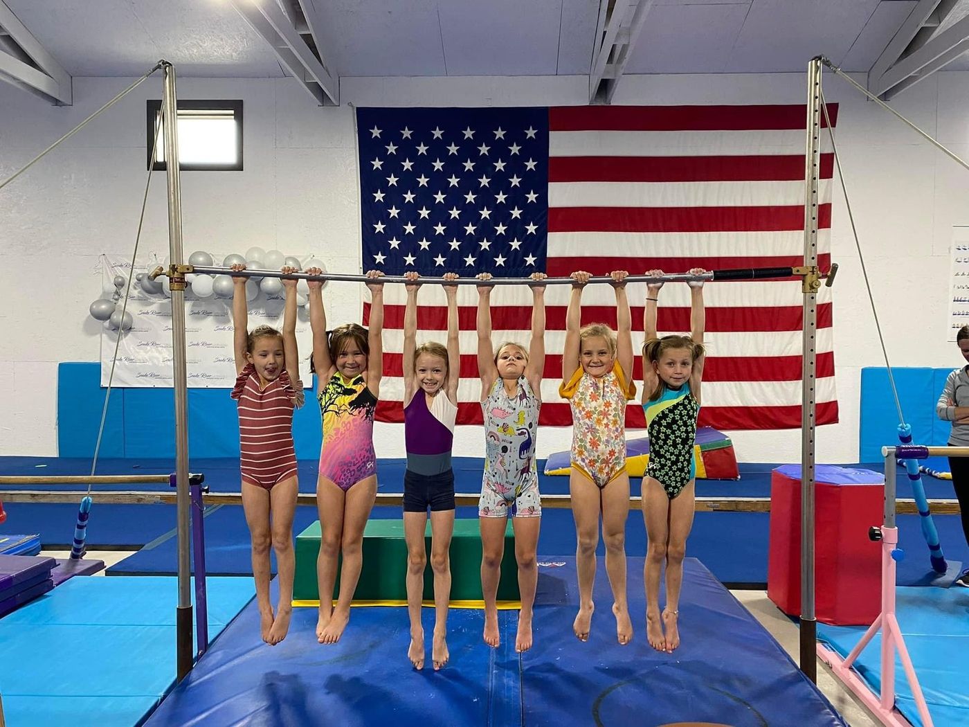 Six young gymnasts hang from the uneven bars in front of a large American flag in the gym.