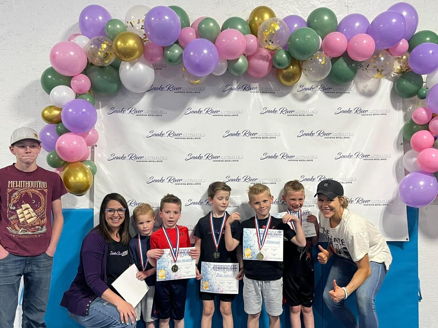 Young boys holding award certificates and medals pose with coaches under a balloon arch at an awards event.