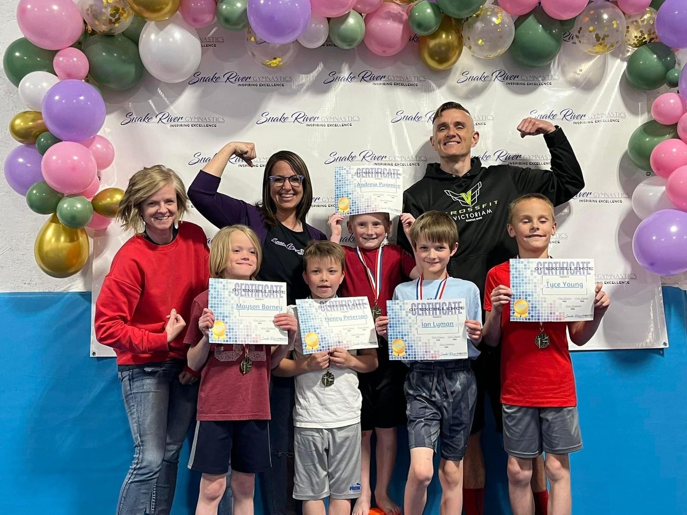 A group of children and adults flex their arms while holding award certificates under a balloon arch.