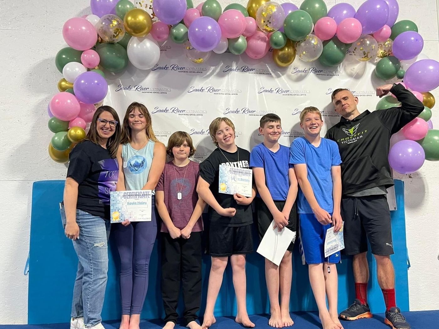 A coach and a group of boys pose with award certificates under a colorful balloon arch.