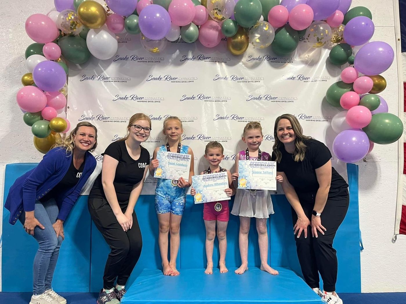 Two coaches kneel beside young gymnasts holding award certificates under a balloon arch.