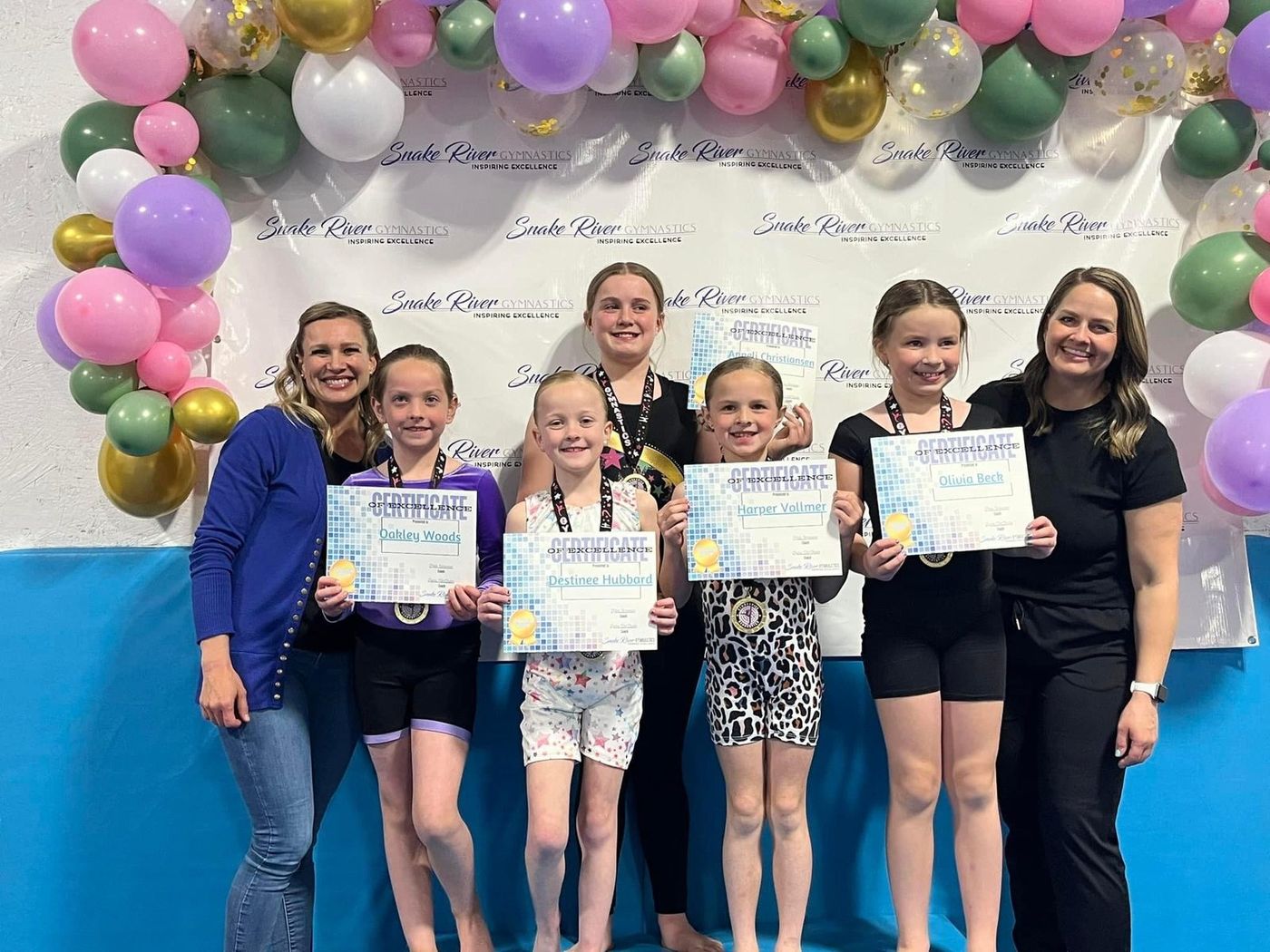 Coaches pose with young gymnasts holding award certificates and medals at an awards ceremony.