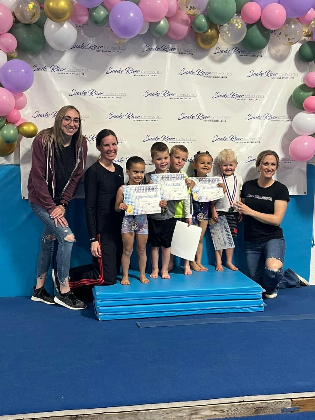 Coaches and young children with medals and certificates pose on a blue mat under a balloon arch.
