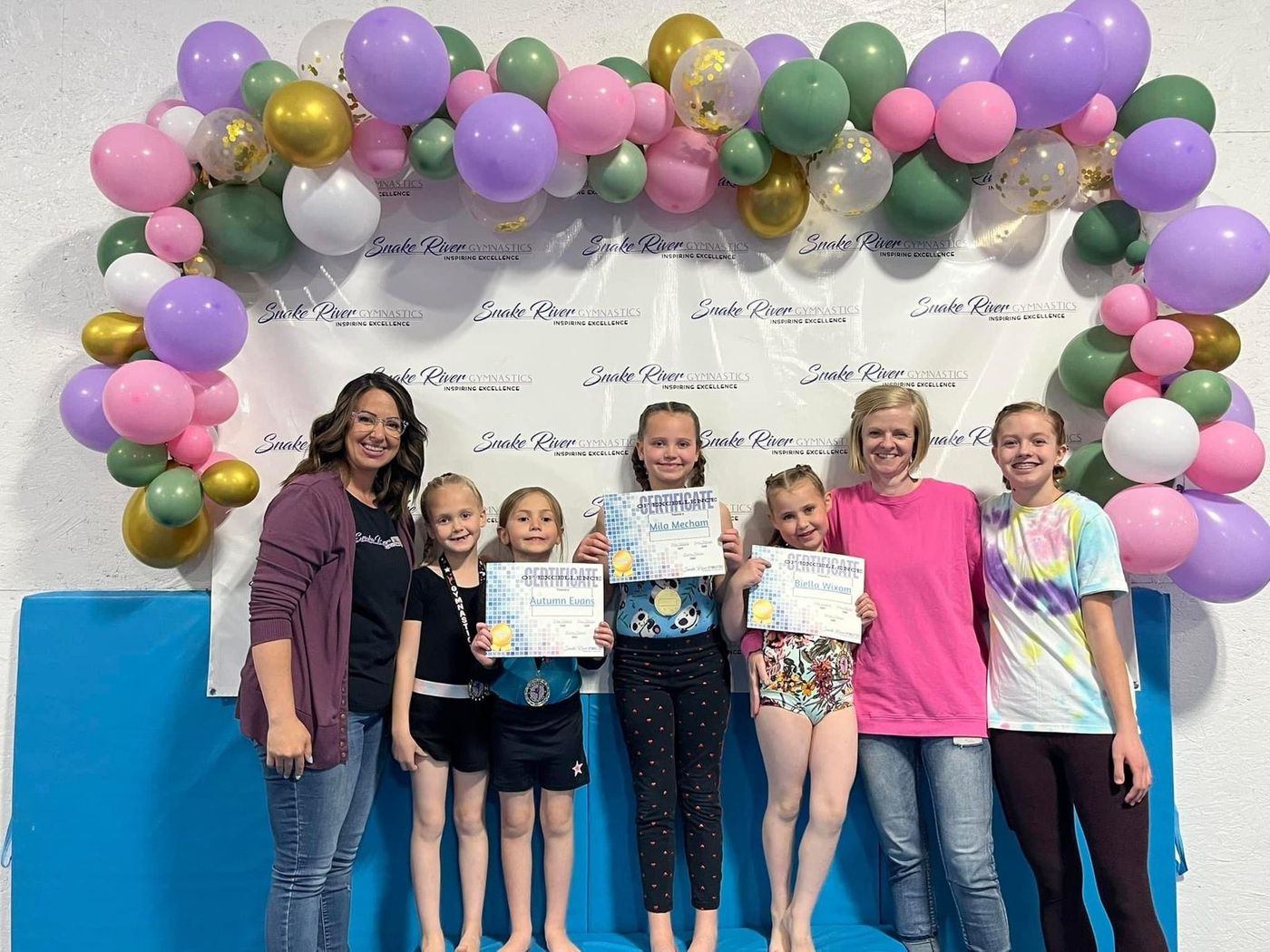 Coaches and young gymnasts holding award certificates pose under a balloon arch in the gym.