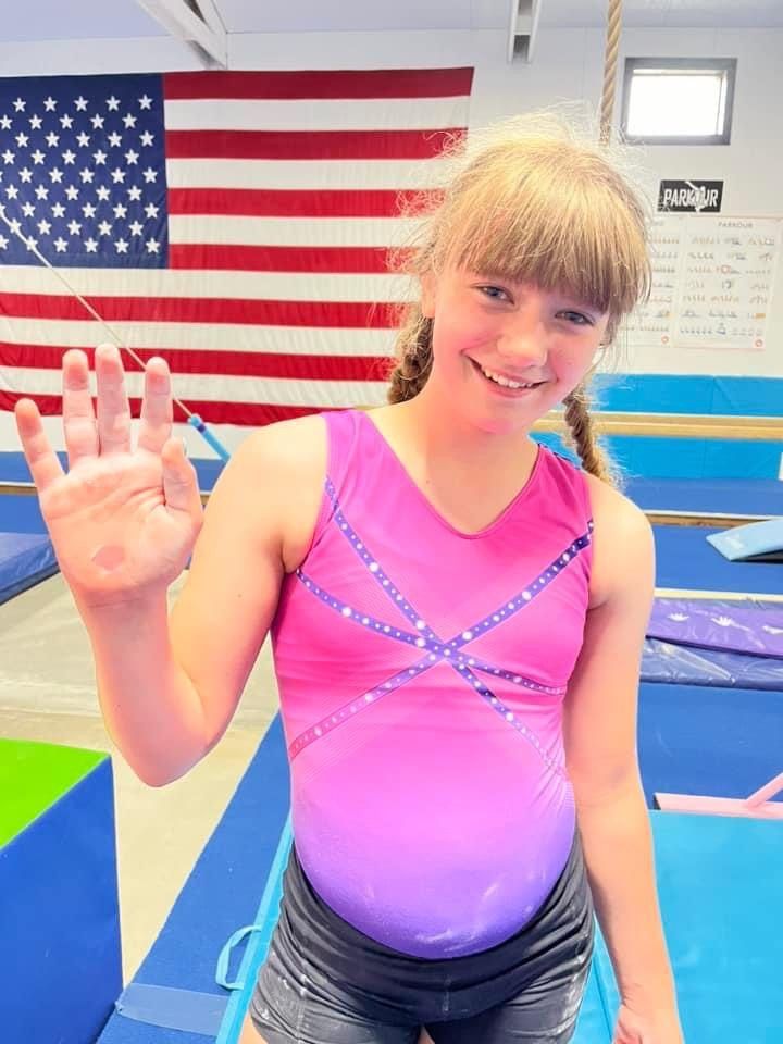 A young gymnast in a pink leotard smiles and waves in front of an American flag in the gym.