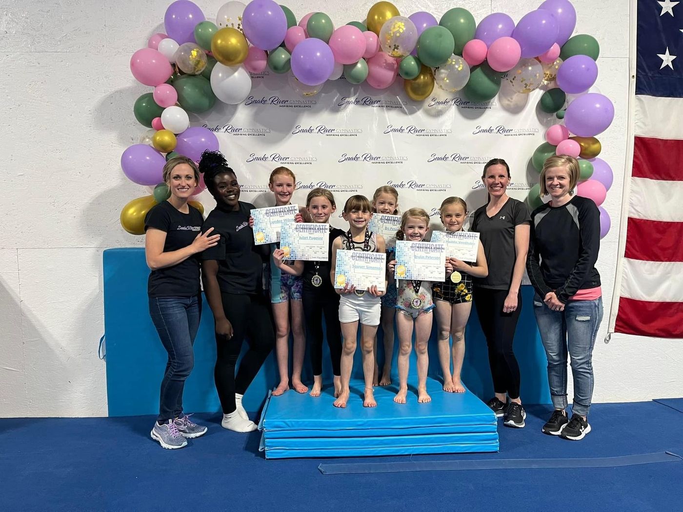 Coaches and young gymnasts hold award certificates and medals on a blue mat under a balloon arch.