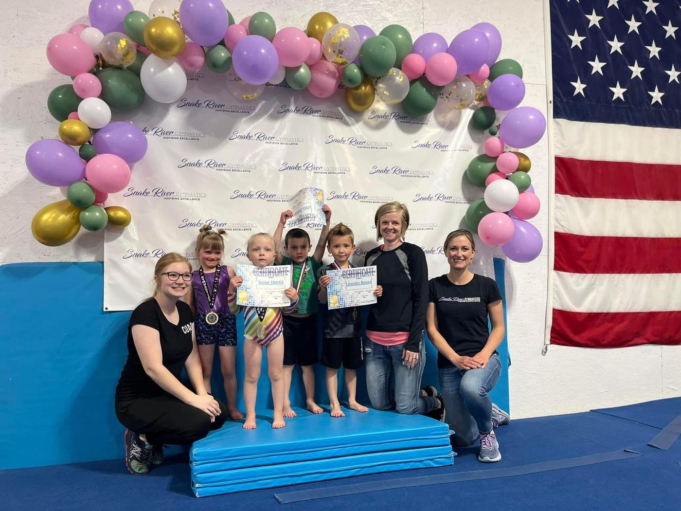 Young children with medals and certificates pose with two coaches under a balloon arch in the gym.