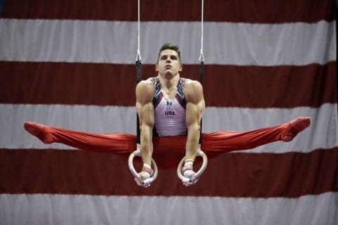 A male gymnast performs an iron-cross hold on the still rings in front of an American flag.
