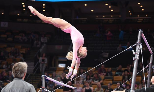 A gymnast in a pink leotard performs a handstand on the uneven bars during competition.