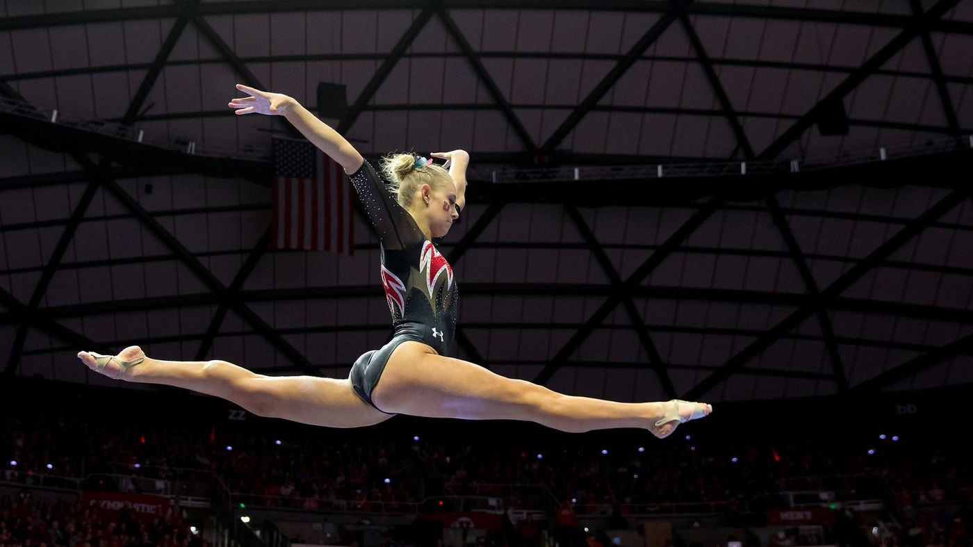 A collegiate gymnast performs a split leap during a floor or beam routine in a packed arena.